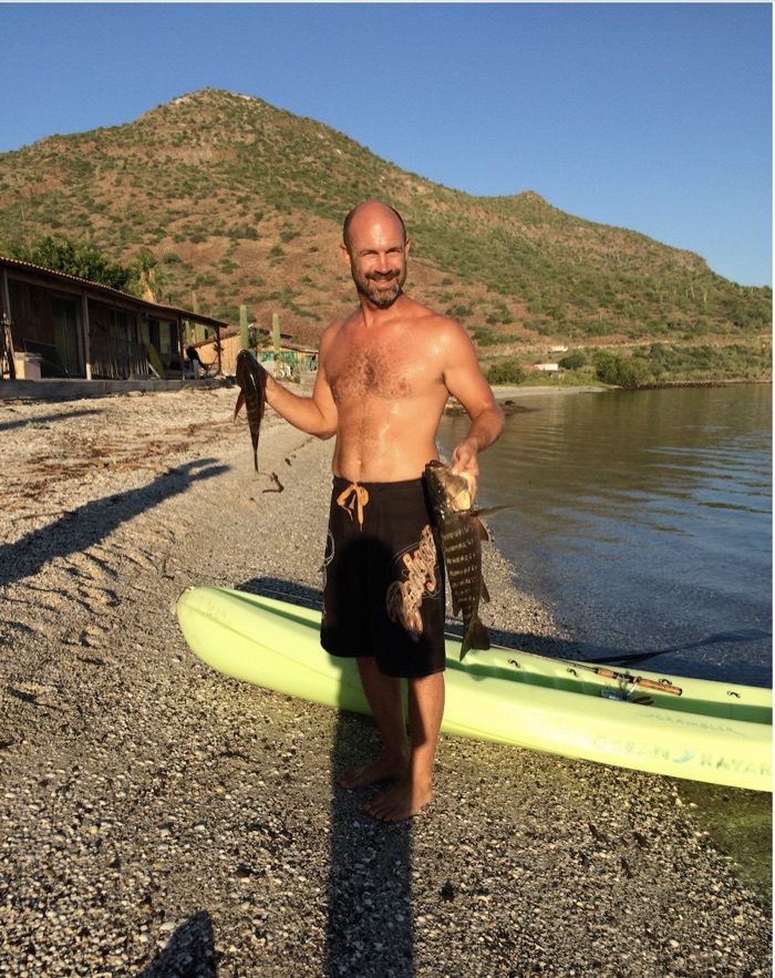 Jed Davis on a beach in Baja, holding two freshly caught fish beside a kayak.