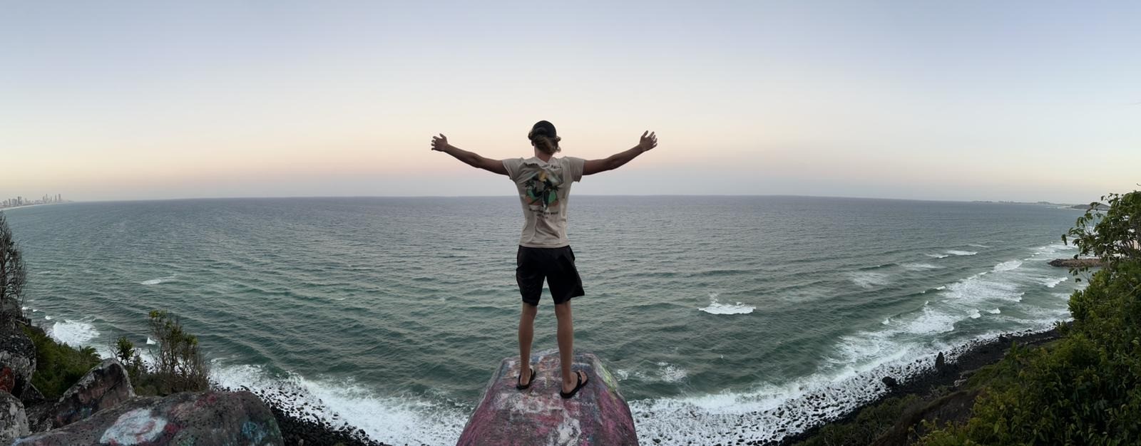 A traveler standing on a cliff with arms spread wide, overlooking the ocean at dusk