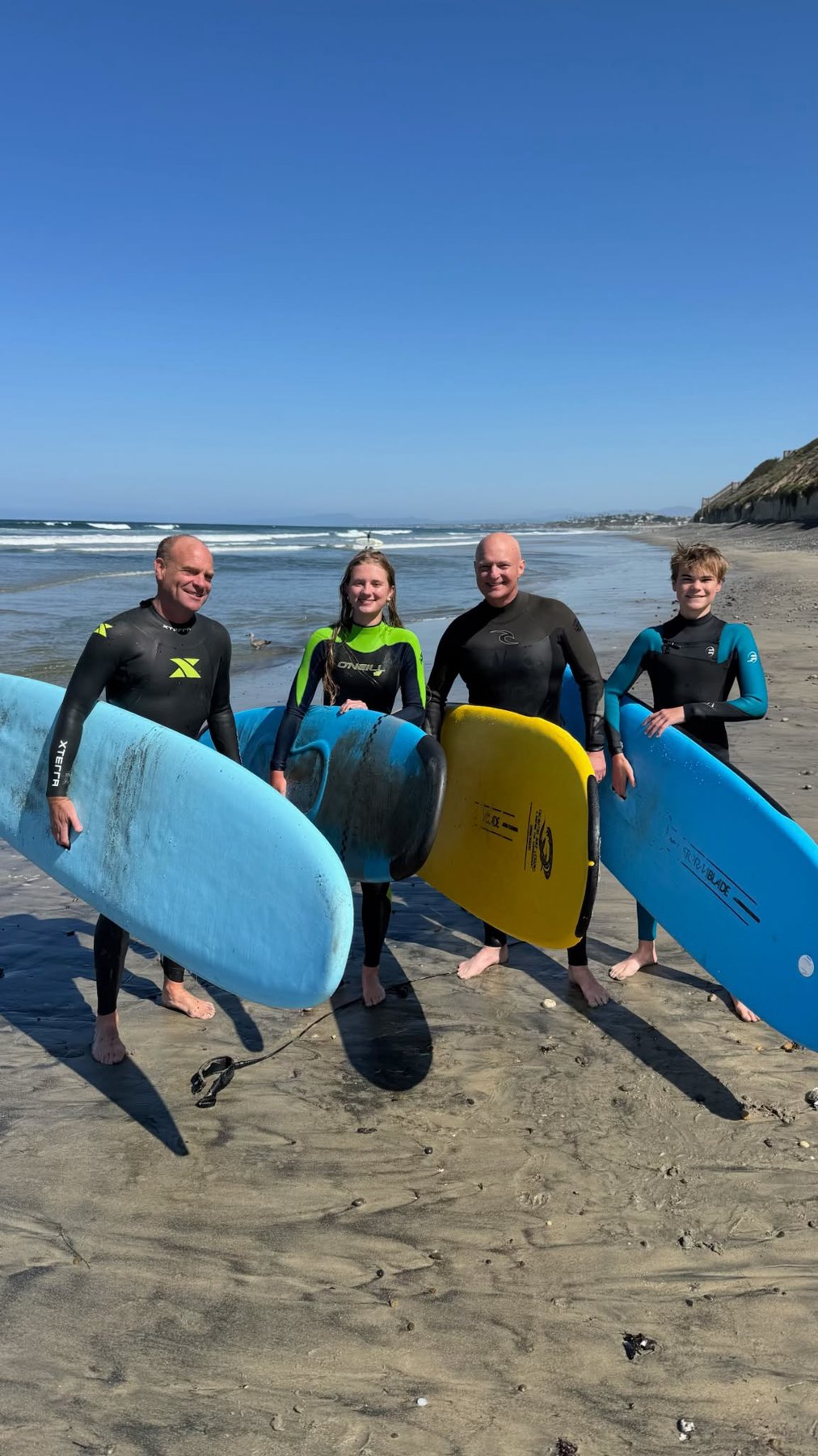 Family of four in wetsuits holding soft-top boards on the sand after a surf lesson