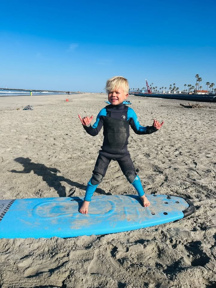 Young surfer in a wetsuit throwing shakas while standing on a blue soft-top on the sand