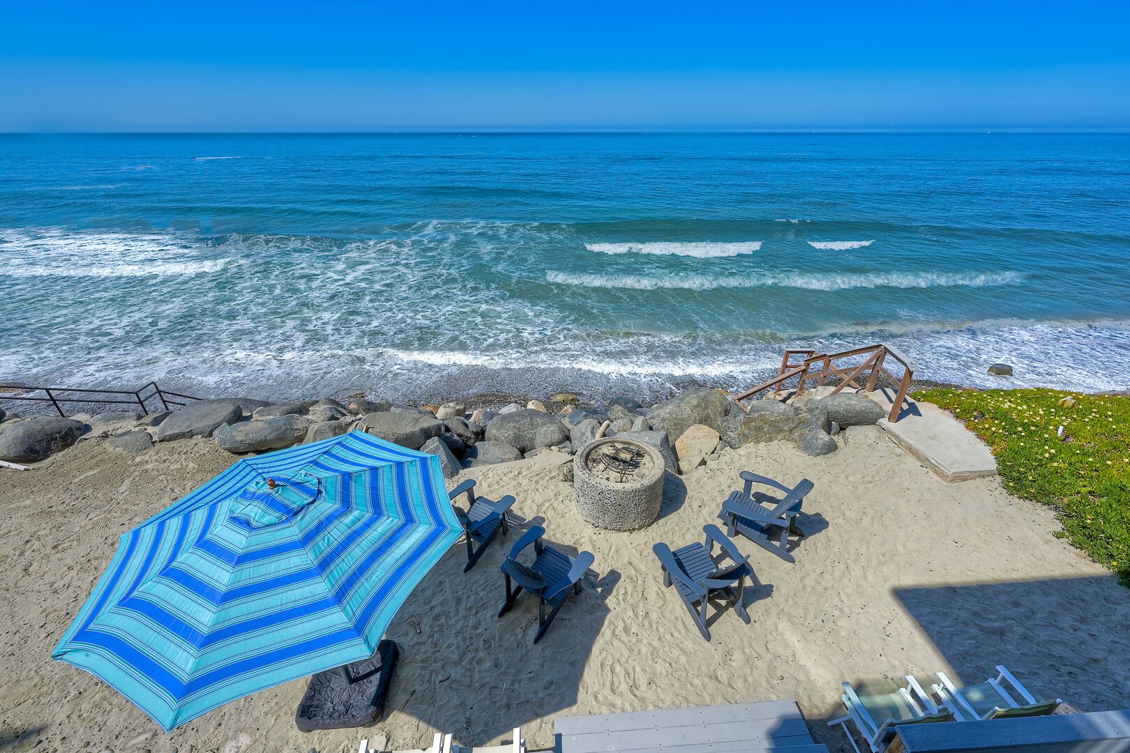 Private sand patio with a fire pit and striped umbrella just above the waves at an Oceanside Haustay home